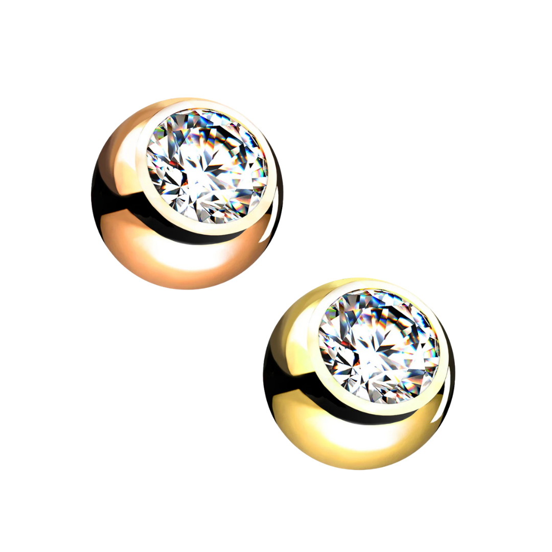 Two replacement piercing barbell balls on a white background, with one featuring a rose gold-plated finish on the left and a yellow gold-plated finish on the right, both inset with a large bezel-set clear cubic zirconia crystal.