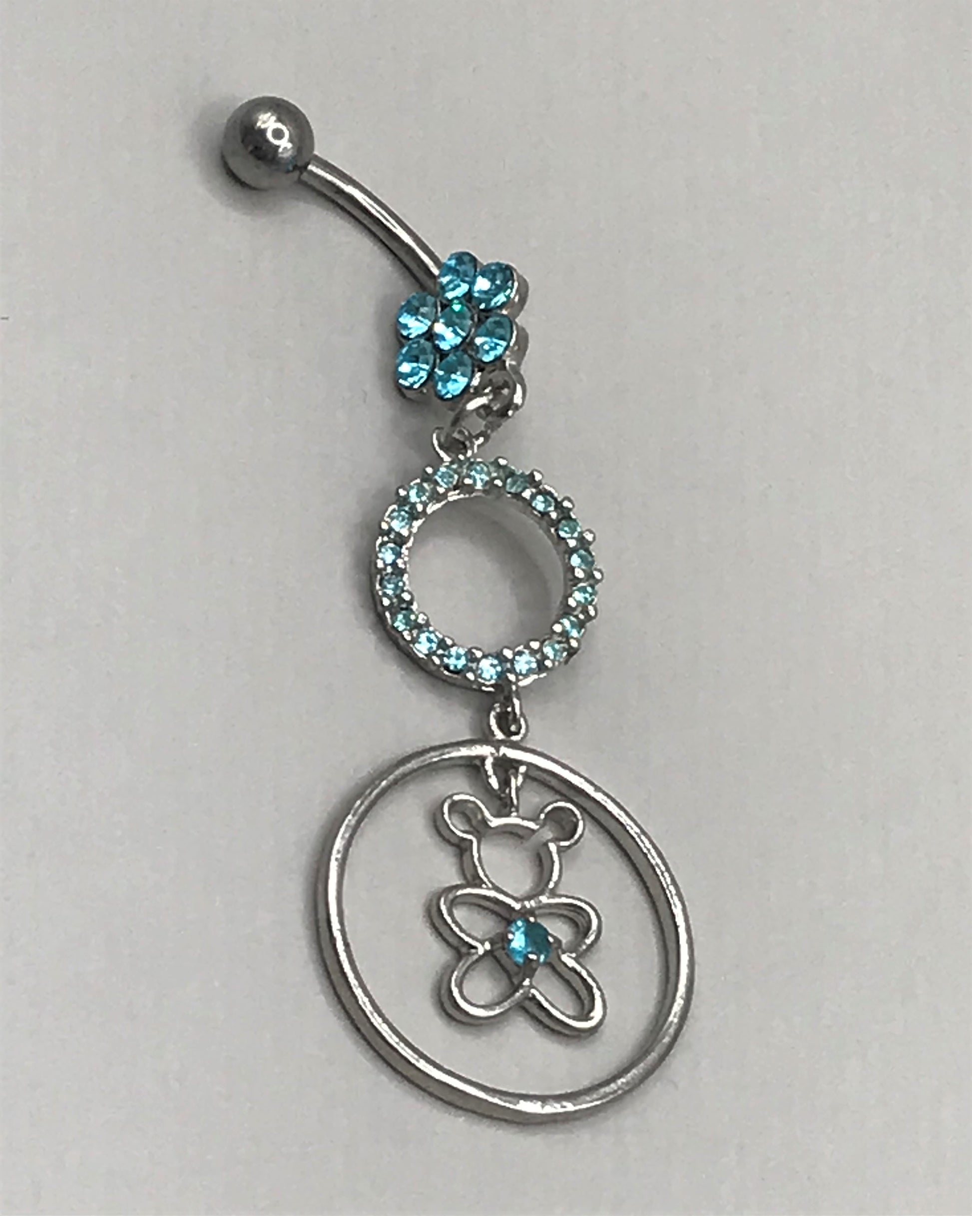 Cute surgical steel belly ring with a bottom gem shaped flower and a dangly small gem covered circle and a bear with a centre gem in a larger circle. Externally Threaded. Shown with light blue gems.