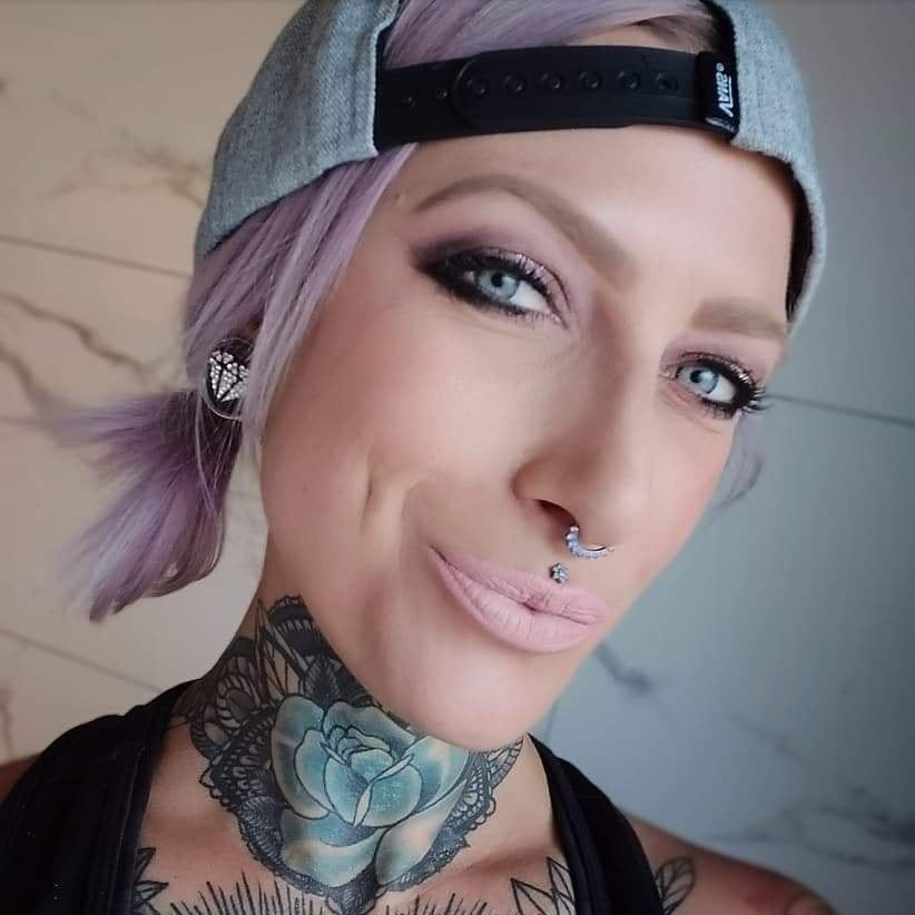 A portrait of a person with lavender hair and a grey cap, featuring a gem-lined silver septum clicker, a sparkling medusa stud, and an ear plug bedazzled with clear gems in a diamond-shaped pattern.