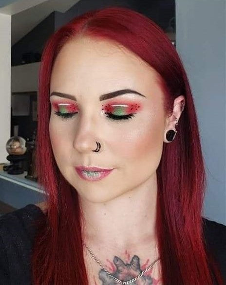 A portrait of a person with vibrant red hair and watermelon-themed eye makeup, featuring a black double nose hoop, black ear plugs, and multiple small silver cartilage piercings.